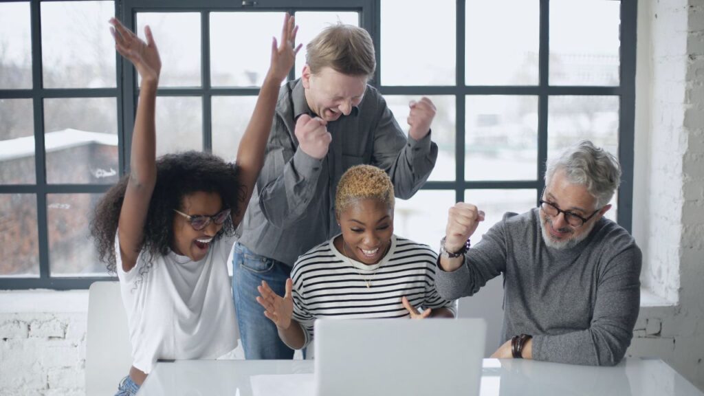 Excited multiracial and multigenerational colleagues enjoying triumph together in front of laptop in office